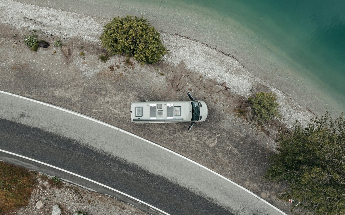 Grå camper van ovenfra, der står på grus ved siden af en sø ved bredden, ved siden af gruset er der en lille vej, og på gruslejet er der tre grønne træer, camperens døre er åbne