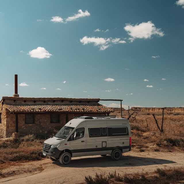 Ljusgrå campervan med svarta designelement står sidan mot en enplans stenbyggnad i ett kargt, ökenliknande landskap under en blå himmel med mycket få små moln