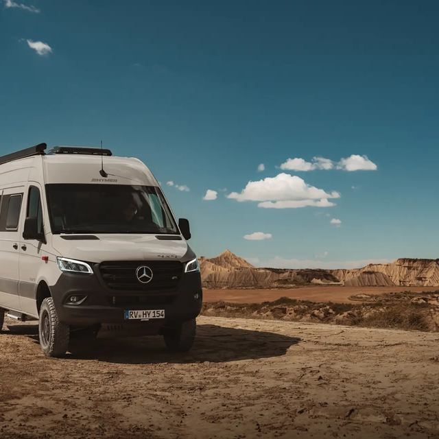 Ljusgrå campervan från HYMER står lätt snett med fronten framåt i ett kargt, ökenliknande landskap under en blå himmel med några vita moln