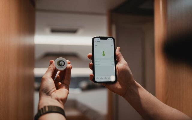 Händerna på en person som håller en temperatursensor i vänster hand och en smartphone som använder Connect-appen i höger hand och mäter den aktuella temperaturen, med husbilens sovområde suddigt i bakgrunden