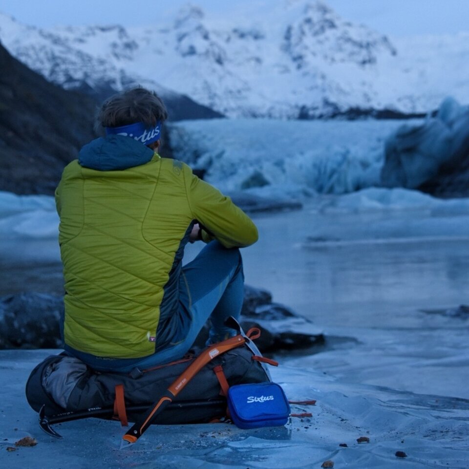 Photo d'un voyage en camping-car en Islande. Un homme en veste verte est assis sur un sac à dos sur une surface glacée, dos au spectateur.