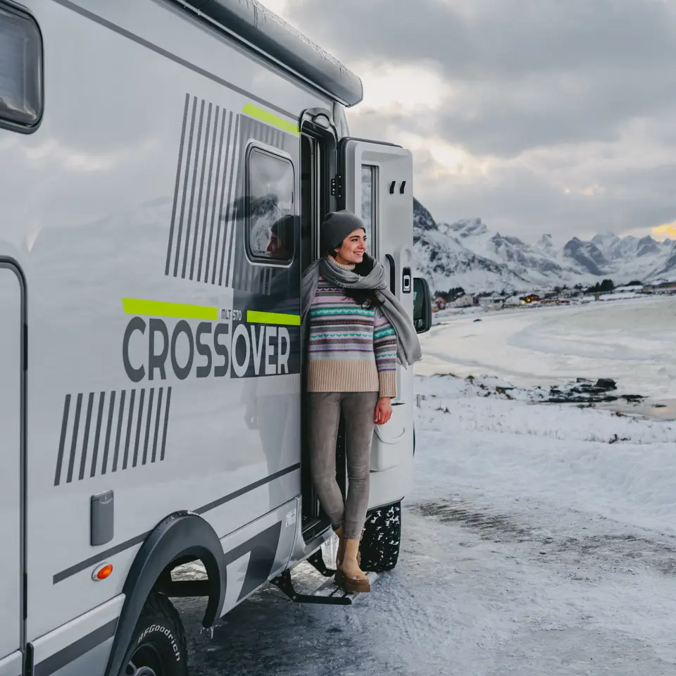 Woman standing at the entrance door of the HYMER ML-T 570 CrossOver, a small village by a sea bay in the background