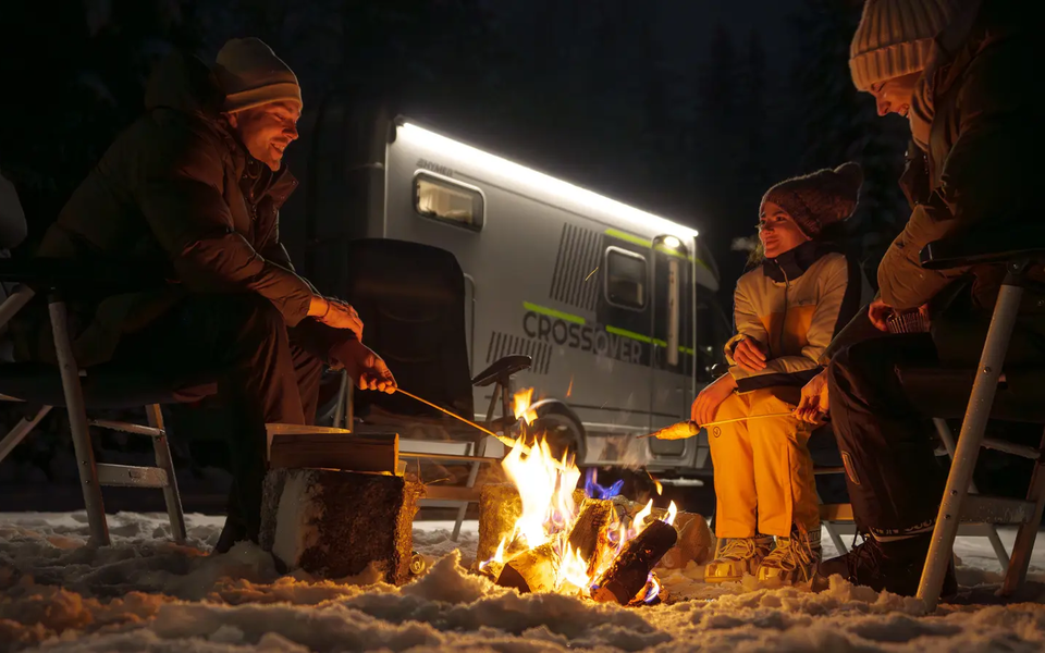 3 people sitting at a campfire making stick bread, the HYMER ML-T 570 CrossOver in the background